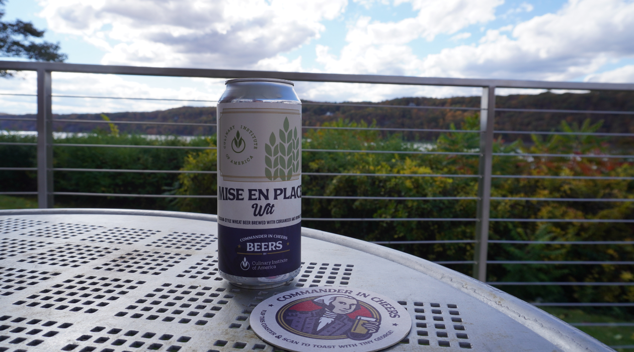 A can of Mise en Place Wit sits on a black table next to Commander in Cheers coaster outside at the patio of The Egg at The Culinary Institute of America in Hyde Park with views of the blue hudson river behind
